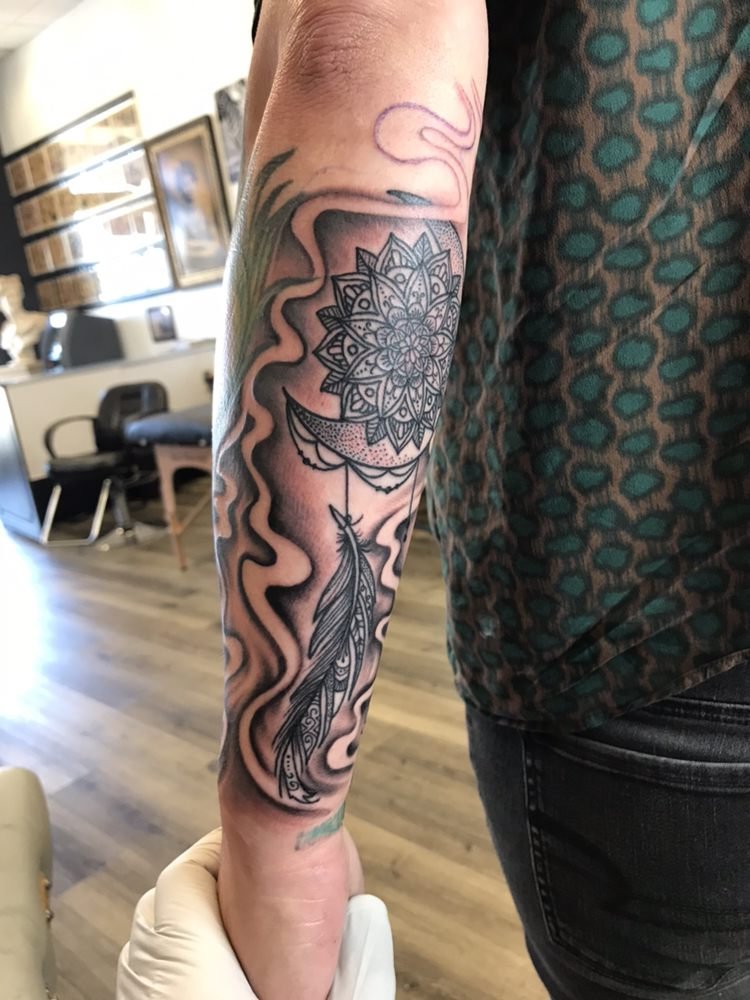 A dotwork mandala over a crescent moon with a hanging feather, set in black smoke on a forearm